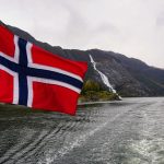 A Norwegian flag flies off the stern of a boat near a shoreline.