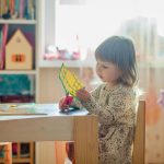 A young girl in a bright classroom engaged in a creative arts and crafts project at a table.