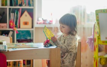 A young girl in a bright classroom engaged in a creative arts and crafts project at a table.